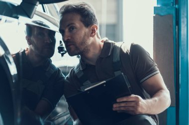 Otomatik mekanik Pano garajda çalışan servis siparişinin tutarak. Onarım Servisi. Siyah arabanın yakınında oturan ve sağlık kontrolü yapma gri üniformalı yakışıklı ciddi beyaz erkeklere