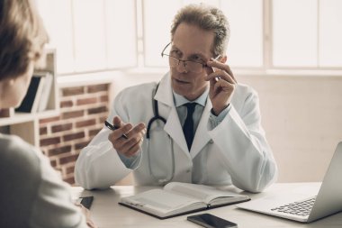 Doktor Ciddi Yüzlü Hasta ile konuşuyor. Dolapta Masada Oturan ve Tanı Hakkında Konuşma Yapan Middleaged Kendinden Emin Sağlık Çalışanı. Tıbbi sorun kavramı