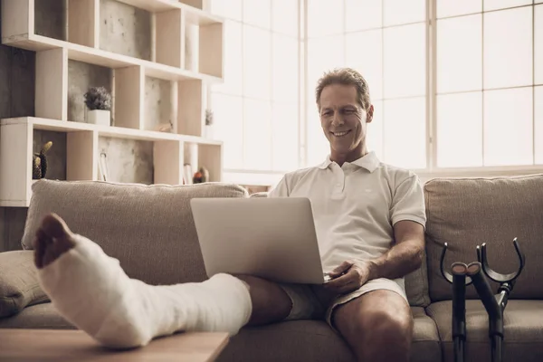 Mann Mit Gebrochenem Bein Sitzt Auf Sofa Und Benutzt Laptop — Stockfoto