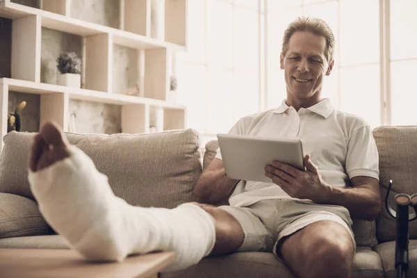 Man With Fractured Leg Sit On Sofa and Use Tablet. Handsome Caucasian Man Broken Leg in Plaster Cast Wearing White Clothes and Posing with Cheerful Expression. Rehabilitation and Health Care Concept
