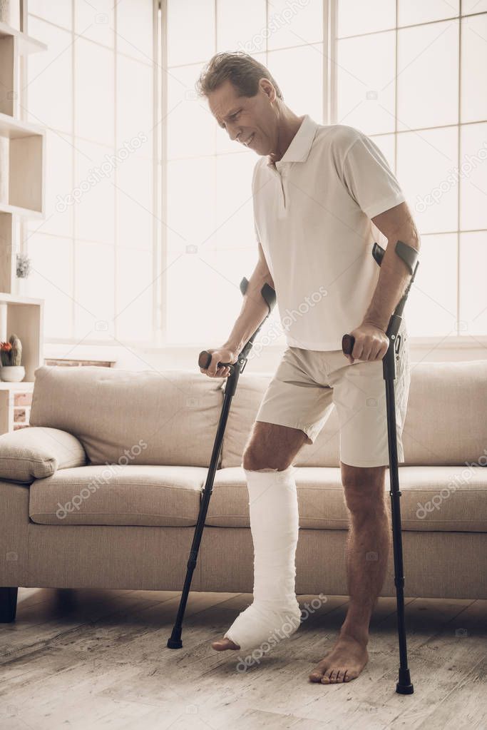 Handsome Man Move Along Room with Crutches. Handsome Caucasian Person with Broken Leg in Plaster Cast Standing near Sofa Indoors with Crutches. Rehabilitation and Health Care Concept