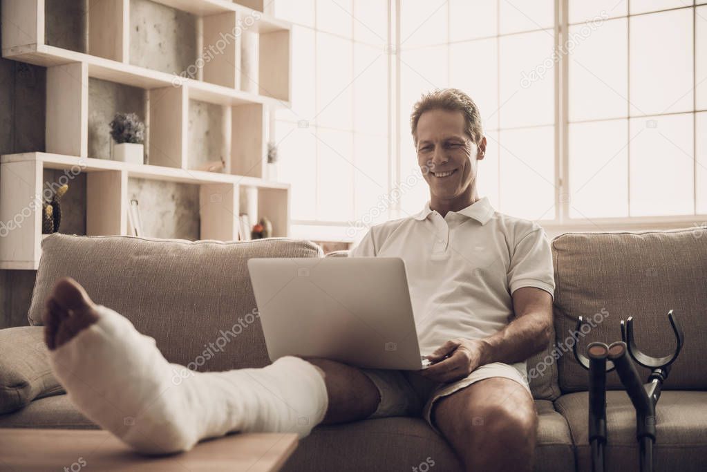 Man With Fractured Leg Sit On Sofa and Use Laptop. Handsome Caucasian Person with Foot in Plaster Cast Wearing White Clothes Posing with Cheerful Expression. Rehabilitation and Health Care Concept