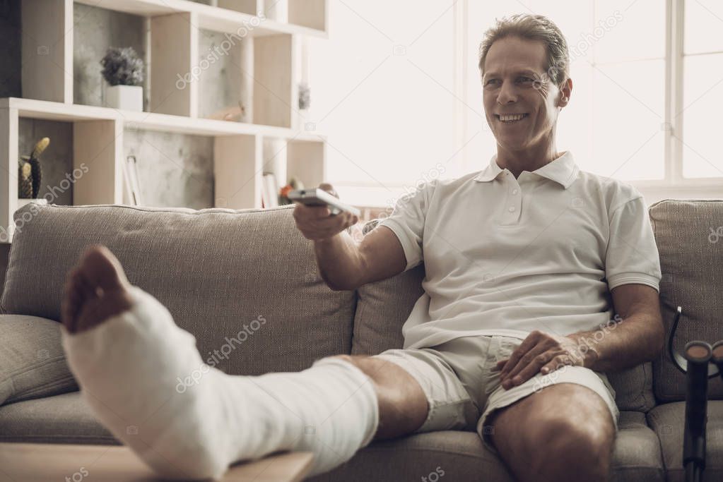 Man With Fractured Leg Sit On Sofa and Watch TV. Handsome Caucasian Person with Foot in Plaster Cast Wearing White Clothes Posing with Cheerful Expression. Rehabilitation and Health Care Concept