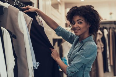 Giyim Mağazasında Gülümseyen Genç Kadın Alışveriş. Mutlu Güzel Siyah Kız Alışveriş Merkezi içinde Butik Satın Almak Için Giysi seçerek. Giyim Mağazası'nda Afro saç modeli ile Çekici Afrikalı Kadın