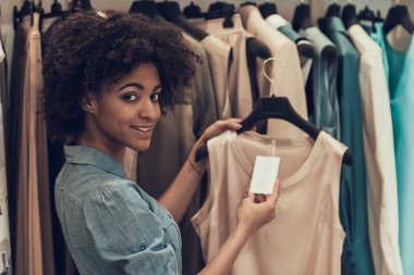 Giyim Mağazasında Gülümseyen Genç Kadın Alışveriş. Mutlu Güzel Siyah Kız Alışveriş Merkezi içinde Butik Satın Almak Için Giysi seçerek. Giyim Mağazası'nda Afro saç modeli ile Çekici Afrikalı Kadın