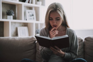 Genç Güzel Kadın Evde Kitap Okuyor. Kanepede Oturan ve Evde Kitap Okuyan Çekici Düşünceli Kız Portresi. Casual kıyafetli Sevimli Kadın Kanepede Kitap Okuyarak Rahatlatıcı