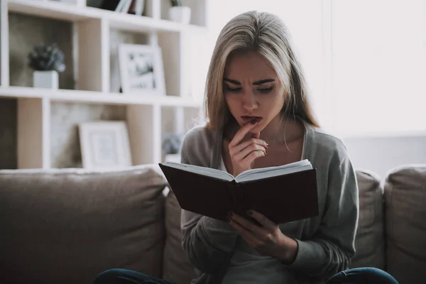 Young Beautiful Woman Reading Book at Home. Portrait of Attractive Thoughtful Girl Sitting on Sofa and Reading Book at Home. Cute Woman in Casual outfit Relaxing by Reading Book on Couch