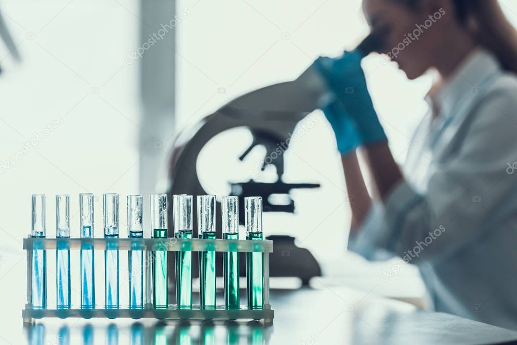 Closeup Chemical Liquid Samples in Flasks in Lab. Female Researcher wearing white Coat sitting at Desk and looking at Samples by using Microscope in Lab. Scientist at Work in Laboratory