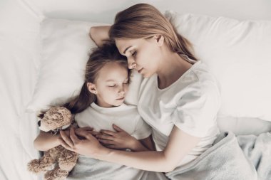 Young Mother and Little Doughter Together in Bed. Beautiful Smiling Woman spending time with Cute Adorable Daughter. Mom and Child holding Teddy bear lying on Bed. Family and Motherhood Concept clipart