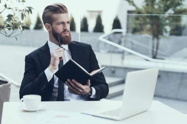 Yakışıklı İşadamı Çalışma Laptop Cafe Outdoors. Not yazıyorum. Sakallı Ciddi Adam Takım Elbise ve Gözlük Giyiyor, Kafede Oturuyor ve Laptop ve İçme Kahvesi ile Çalışıyor. İş Kavramı.