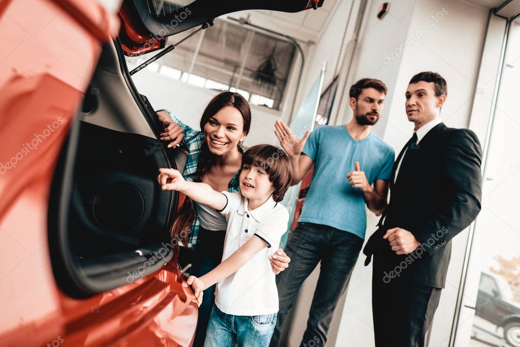 Happy Young Family Are Choosing A New Auto In Showroom. Dialogue With Dealer. Cheerful Customer. Look Inside The Trunk. Make A Decision. Good Offer. Buyer And Seller. Business Trade.