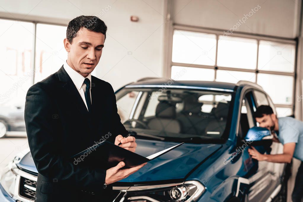 Car Dealer Camera Posing With A Buyer On Background. Cheerful Customer. Automobile Salon. Make A Decision. End Of A Deal. Good Offer. New Buying. Business Trade. Confident Seller. Purchase Order.