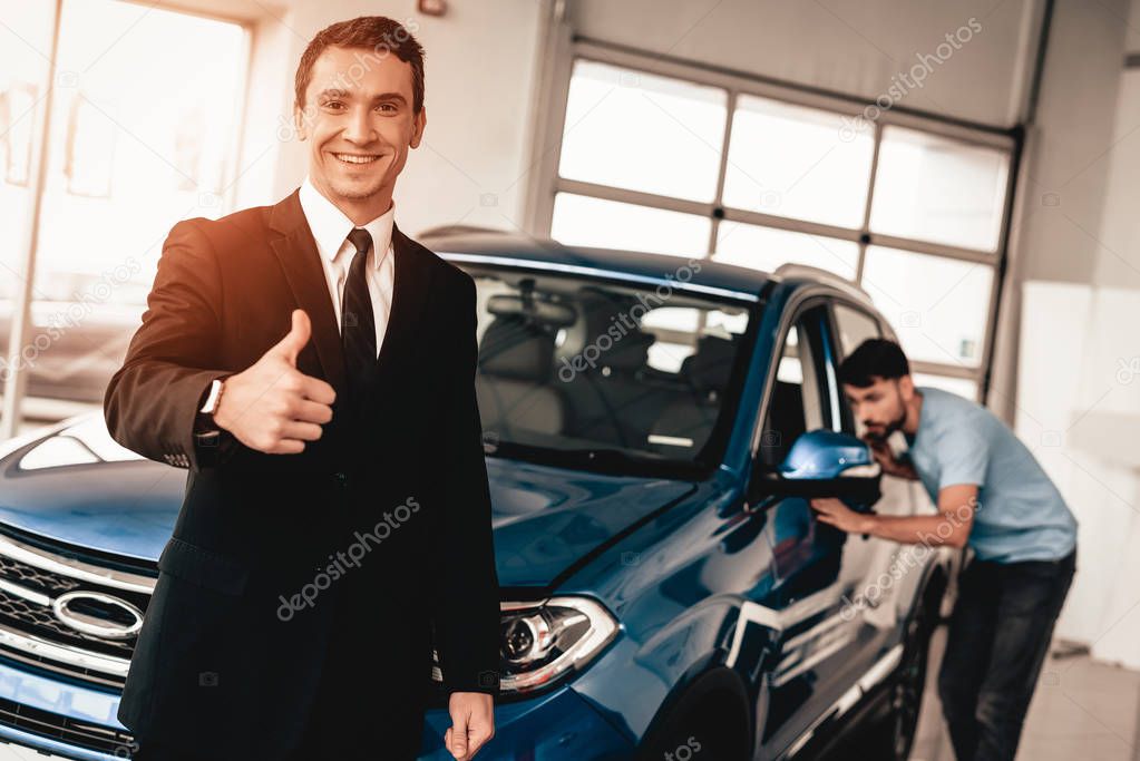 Car Dealer Camera Posing With A Buyer On Background. Cheerful Customer. Automobile Salon. Make A Decision. End Of A Deal. Good Offer. New Buying. Business Trade. Confident Seller. Thumb Up.