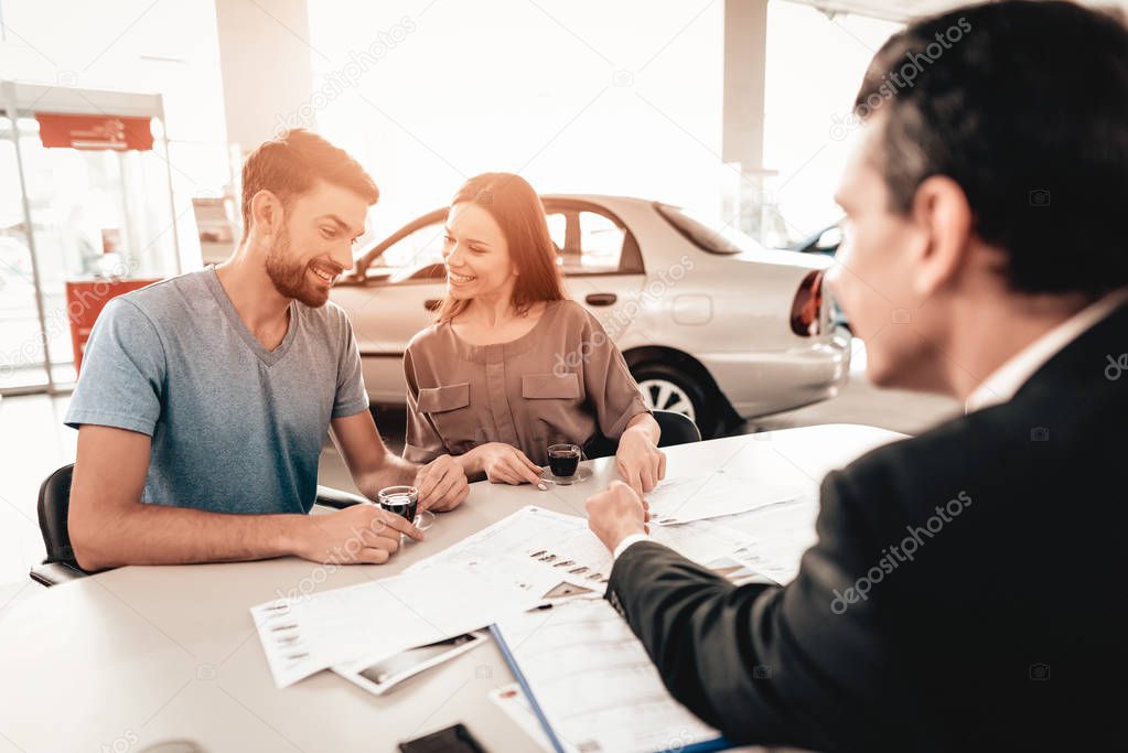 Happy Young Family Are Choosing A New Car In Showroom. Dialogue With Dealer. Cheerful Customer. Automobile Salon. Make A Decision. Cup Of Coffee. End Of A Deal. Good Offer. Buyer And Seller.