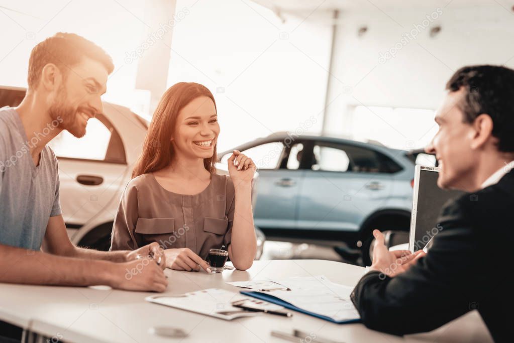 Happy Young Family Are Choosing A New Car In Showroom. Dialogue With Dealer. Cheerful Customer. Automobile Salon. Make A Decision. Cup Of Coffee. End Of A Deal. Good Offer. Information On Tablet.