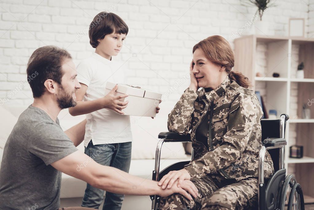 Woman Veteran In Wheelchair Homecoming Concept. Family Meeting. Son And Husband. Box With A Gift. Camouflage Uniform. Child Hanging. Feelings Showing. Patriotic Comeback. Paralyzed Soldier.