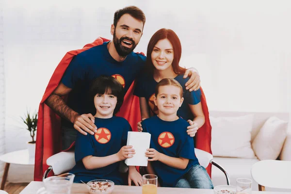 年轻的家庭在超级英雄西装 晚餐的概念 T恤衫和雨衣 明亮的客房 在一起休息 果汁和谷物 准备好 健康的生活方式 有父母的孩子拿着盒子 — 图库照片