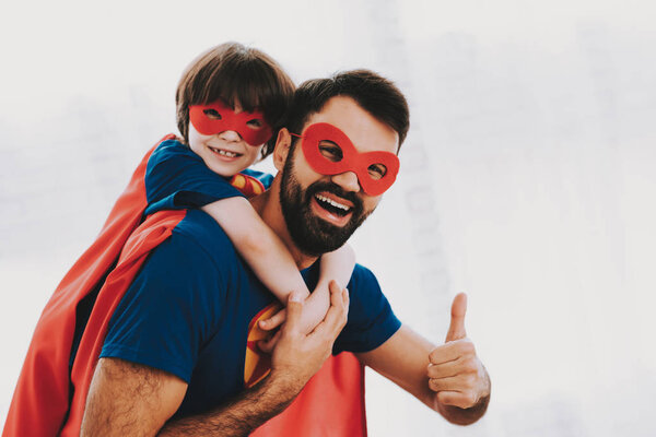 Father And Son. Red And Blue Superhero Suits. Masks And Raincoats. Posing In A Bright Room. Young Happy Family Holiday Concept. Resting Together. Save The World. Get Ready. Thumbs Up.