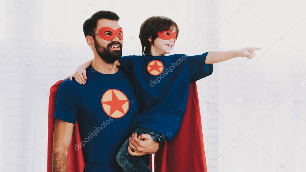 Father And Son. Red And Blue Superhero Suits. Masks And Raincoats. Posing In A Bright Room. Young Happy Family Holiday Concept. Resting Together. Save The World. Get Ready. Strong And Powerful.