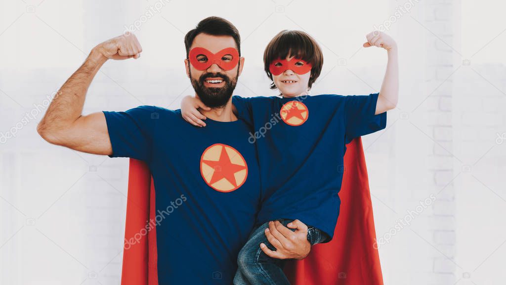 Father And Son. Red And Blue Superhero Suits. Masks And Raincoats. Posing In A Bright Room. Young Happy Family Holiday Concept. Resting Together. Save The World. Get Ready. Strong And Powerful.