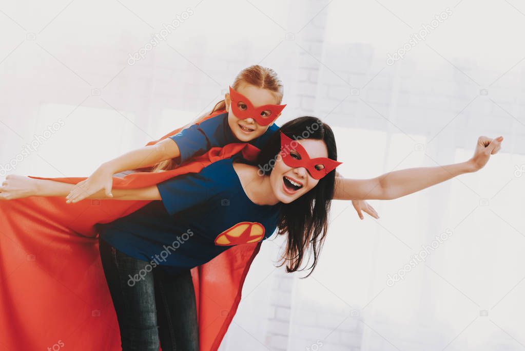 Mother And Daughter. Red And Blue Superhero Suits. Masks And Raincoats. Posing In A Bright Room. Young Happy Family Holiday Concept. Resting Together. Get Ready. Strong And Powerful.
