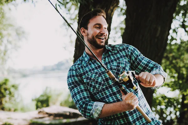 Man in Checkered Shirt Fishing on River in Summer. Relaxing Outdoor. Sitting Man. Man near Lake. Fishing Rod in Hands. Sports in Summer. Man in Shirt. Bearded Guy. Weekend on River. Young Fisherman.