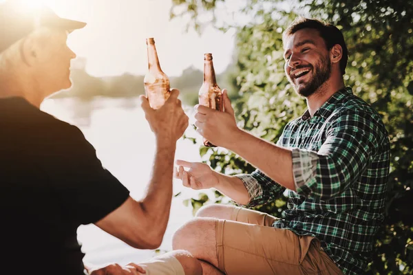 三夏の川の近くのビールを飲んで笑顔男性シャツを着た男性 ひげを生やした男 川の週末 若い男はリラックス屋外 湖の近くに座って男男 アルコールを飲みます 瓶ビール — ストック写真