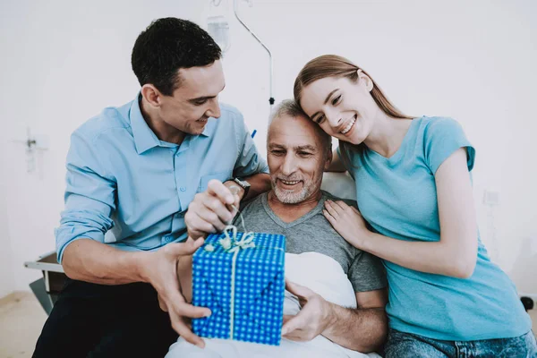 Gift for Old Man in Clinic. Young Family give Old Man Gift. Happy Family. Good Mood in Family. Family in Clinic. Old Man and Family in Hospital. Health Man and Family in Clinic. Care about Old Man.