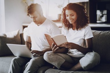 Dizüstü bilgisayar ve kitap kanepede oturan bir çift gördüm. Sabah eğlence. Mola verebilir. Ekrana bak. Kanepenin üzerinde tüyler ürpertici. Ev giysileri. Tatil dinlenme. Birlikte gülümseyen. Eller klavye.