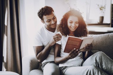 Adam kitap kız kanepede okuma asılı duruyor. Sabah eğlence. Mola verebilir. Ev giysileri. Tatil dinlenme. Birlikte gülümseyen. Mutlu genç Afro Amerikan çift. Güneşli bir gün. Birbirimizi seviyoruz.