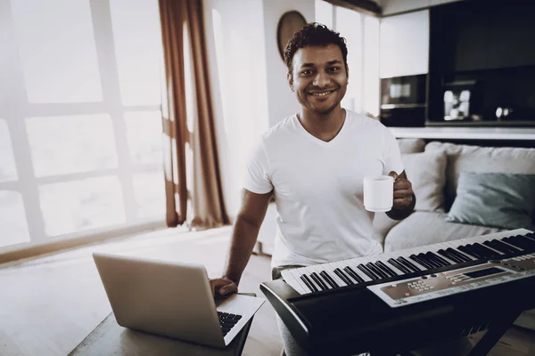 Young Man Drinks Coffee Near Synthesizer At Home. Happy Songwriter. Morning Leisure. Digital Format Sound Record. Working Musician. Musical Hobby. Cup Of Coffee. Working With Laptop.