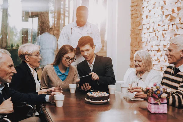 Happy Together. Parents. Children. Grandparents. Tablet. Talk. Good Mood. Doctor at Head. Young and Old People. Sit Together. Table. Room. Nursing Home. Drink Different Drinks. Visit. Happy Family.