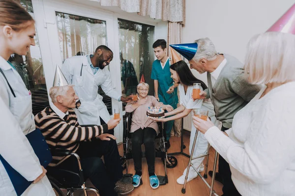 Group. Young and Old People. Nursing Home. Congratulate. Elderly Woman. Birthday. Very Surprised. Very Happy. Glasses of Drinks. Nurse Hold Cake. Birthday Party. Celebration. Gifts. Balloons. Guests.