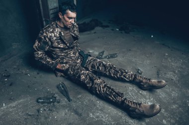 Adam savaş barınak av tüfeği ile oturuyor. Kamuflaj üniforma. Askeri eylemler. Savaş Hideaway kavramı. Şişe alkol ile. İşgal sorunu. Depresif asker. Hayal kırıklığına kahraman.