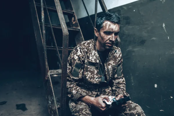 Man Is Sitting In War Shelter With Shotgun. Camouflage Uniform. Military Actions. War Hideaway Concept. Occupation Problem. Depressed Soldier. Disappointed Hero. Posttraumatic Syndrome.