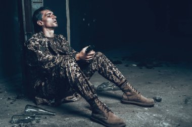 Adam savaş barınak av tüfeği ile oturuyor. Kamuflaj üniforma. Askeri eylemler. Savaş Hideaway kavramı. Şişe alkol ile. İşgal sorunu. Depresif asker. Hayal kırıklığına kahraman.