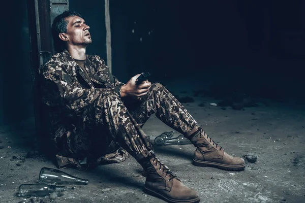 Man Is Sitting In War Shelter With Shotgun. Camouflage Uniform. Military Actions. War Hideaway Concept. Bottle With Alcohol. Occupation Problem. Depressed Soldier. Disappointed Hero.