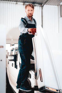 Kızıl saçlı makinist tekerlek üzerinde duruyor. Açık Hood.Professional üniforma. Kendine güvenen Mühendisliği uzmanı bakışları. Koruyucu eldiven. Garajda çalışıyor. Genç tamirci kavramı poz. Büyük Suv.