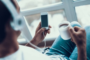 Engelli adam müzik ve içecekler kahve için dinler. Portre, kişi oturur pencere yakınındaki tekerlekli sandalyede fincan çay elde ile rahat kıyafetler giyer und kullanan akıllı telefon ve kulaklık.