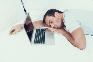 Adam Asleep dizüstü bilgisayarda çalışırken yatağa düştü. Beyaz çarşaflar hafif renkli tişört giyen ile büyük yatakta açılan not defteri yakınındaki yatan yorgun yakışıklı kahverengi saçlı genç insan portresi