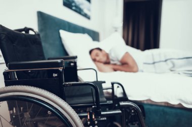 Siyah tekerlekli sandalye yakınındaki Engelli kişi uyur. Ön plan siyah Modern geçersiz sandalyeye closeup. Parlak yatak beyaz keten ile büyük yatakta yatan özürlü genç insan