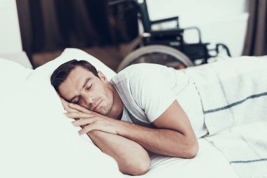 Alarm beyaz keten ile yatakta yakınındaki kişi uyur. Eller başının altına tutmak ve beyaz T-shirt giyen artalanları üzerinde bulanık tekerlekli sandalye ile yatak odasında yatan yakışıklı genç adam. Sabah kavramı