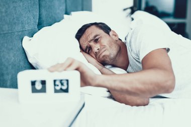 Man Hardly Awakes and Pulls Alarm Off in Morning. Handsome Young Person in Bad Mood Lying in Bedroom in Bed With White Linens Near Alarm Clock and Wearing White T-shirt. Morning Concept clipart