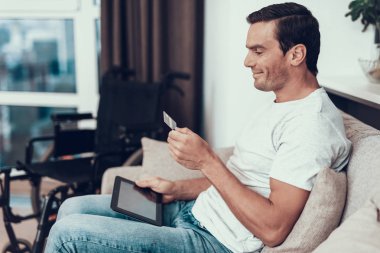 Engelli kişi kredi kartı tutan ve Tablet kullanır. Çevre tekerlekli sandalye Online mu kanepede oturan adam gülümseyen yakışıklı ile alışveriş Modern oturma odası yüz ifadesinde memnun