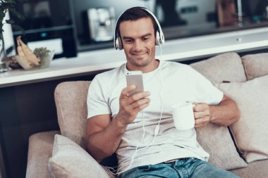 Adam müzik kulaklık ve içecek çay dinler. Akıllı telefonlar kulaklık kullanarak ekrana bakarak kanepede otururken yakışıklı genç gülümseyen portresi. Erkek tutar fincan kahve elinde.