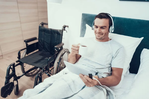 Man Watch Video in Pad in Headphones and Hold Cup. Portrait of Happy Handsome Invalid Lies in White Bed Near Modern Black Wheelchair and Looks at Tablet Screen and Drinks Coffee or Tea