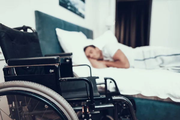 Disabled Person Sleeps Near Black Wheelchair. Closeup of Black Modern Invalid Chair on Foreground. Handicapped Young Person Lying in Large Bed With White Linens in Bright Bedroom