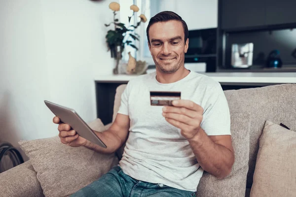 Cheerful Person Holds Credit Card and Uses Tablet. Handsome Young Smiling Man Sitting on Bright Comfortable Couch Does Online Shopping in Modern Living Room Showing Satisfied Face Expression
