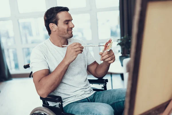 Handicapped Man in Wheelchair Painting Picture. Smiling Handsome Happy Caucasian Person Sitting Opposite Easel in Living Room Wearing Casual Clothes Holding Paintbrush and Palette