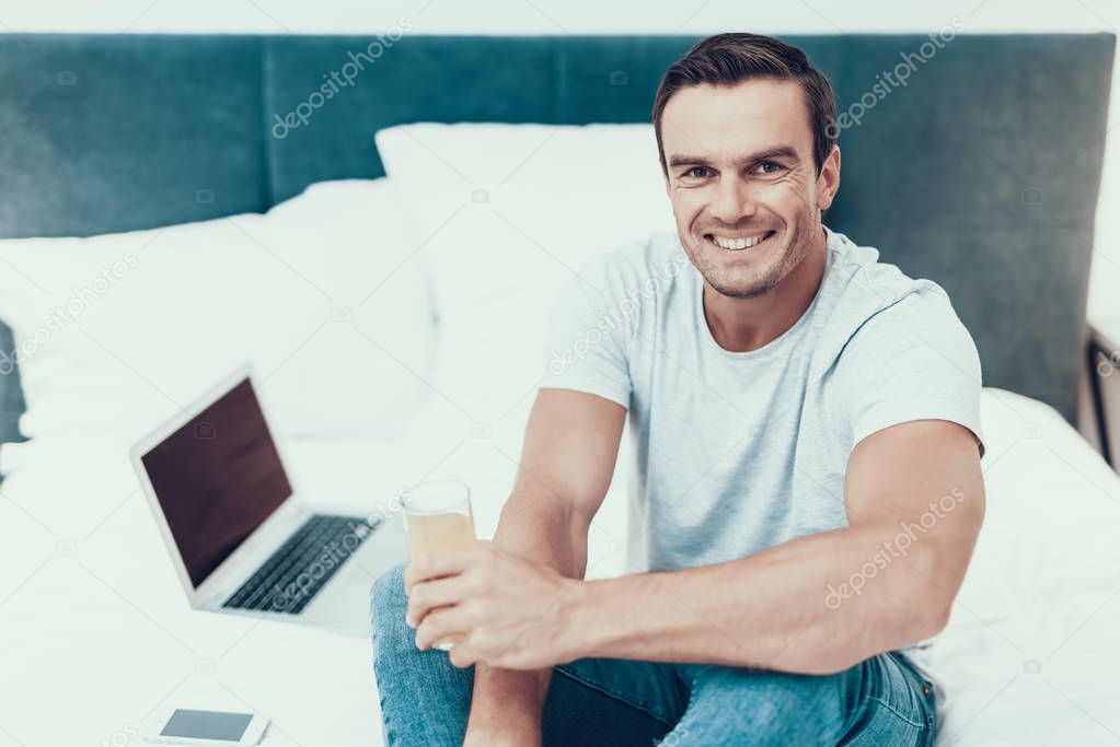 Man Sits on Bed with Laptop and Drinks Juice. Portrait of Caucasian Happy Person Wears Casual Clothes Works on Laptop, Has Breakfast and Looks at Camera in Modern Apartment